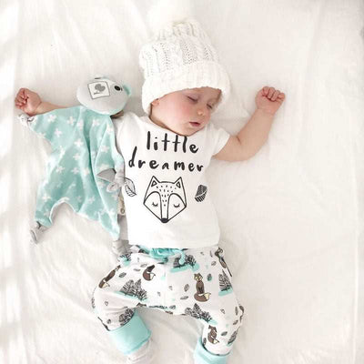 Newborn baby in comfortable cotton clothes set with "little dreamer" T-shirt and pants, lying next to a plush toy.