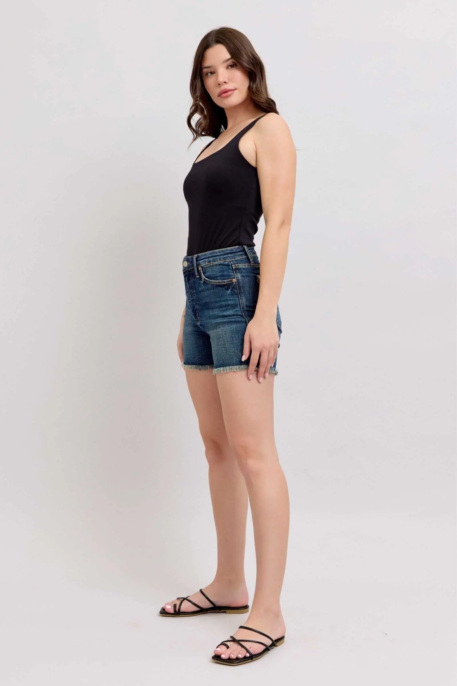 Judy Blue Tummy Control fray hem denim shorts with shield back pockets, worn by woman in black tank top and sandals.